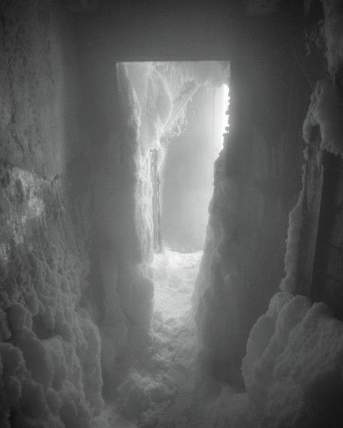 a black and white photograph shows a doorway leading into a snowy tunnel. the walls are caked in snow and ice, giving the impression of a blizzard. the scene is monochromatic, enhancing the cold and stark atmosphere.