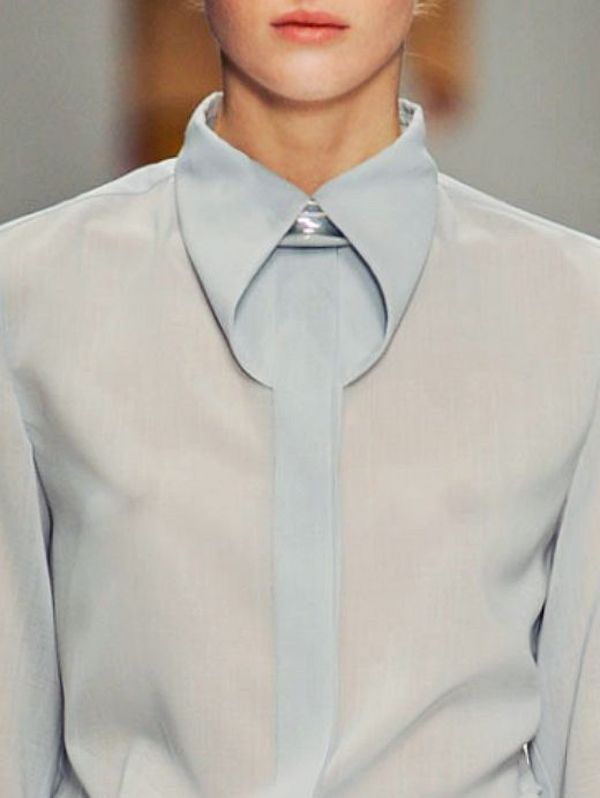 closeup view of a woman's upper body, showcasing a light blue, sheer blouse with a distinctive, oversized collar. the collar is folded and points downward, creating a modern and elegant look. the woman's face is partially visible, with a neutral expression. the overall image suggests a highfashion setting, possibly a runway or photoshoot. the focus is on the unique design of the shirt's collar and the subtle sheen of the fabric.