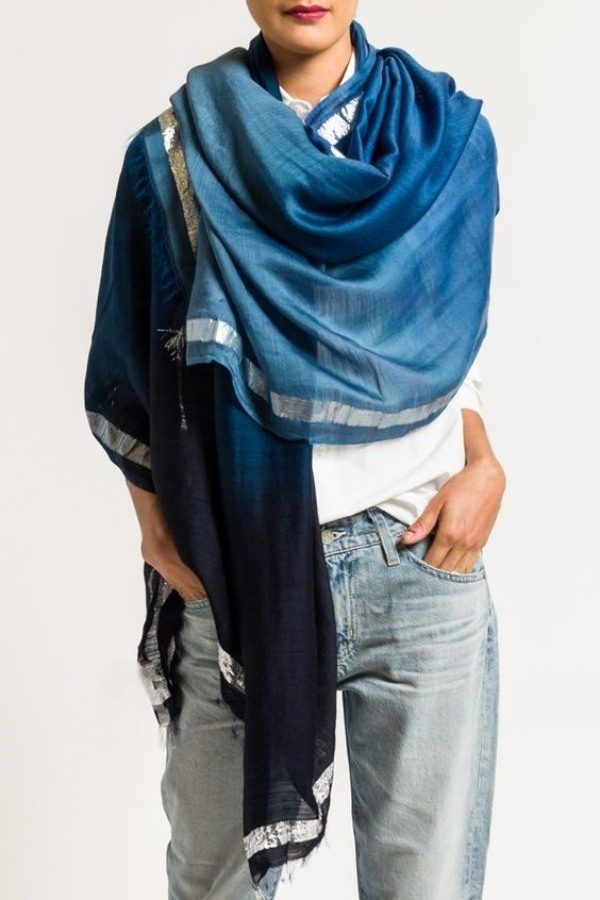 a woman is wearing a large blue scarf with a silver trim, jeans, and a white top. the scarf is draped around her neck and shoulders.