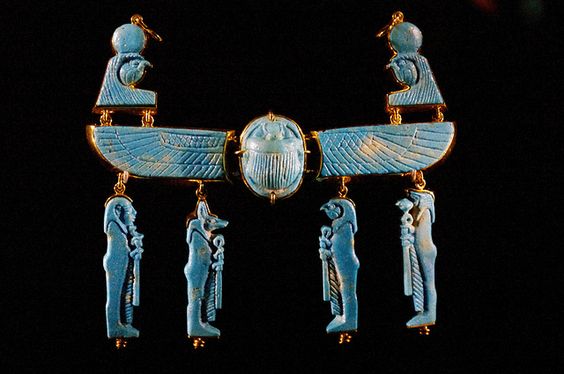 this striking piece is an ancient egyptian necklace made of gold and turquoise. the centerpiece is a large scarab beetle, flanked by outstretched wings. hanging below the wings are four figures, two on each side. the figures represent egyptian gods and goddesses, showcasing the intricate craftsmanship and religious significance of the piece. the necklace is set against a black background, illuminated by spotlights that highlight its beauty and detail.