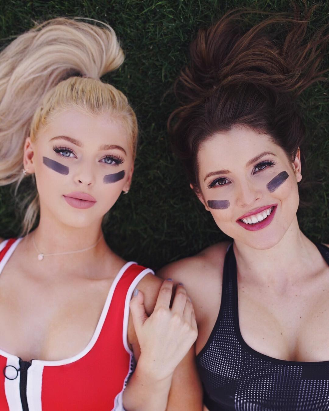 two women are lying on the grass, both wearing eye black. the woman on the left has blonde hair in a high ponytail and is wearing a red and white top and a necklace. the woman on the right has brown hair in a messy bun and is wearing a black and gray sports bra. both women are smiling and wearing lipstick.