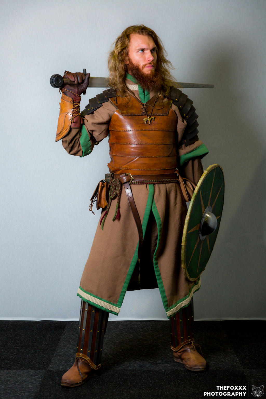 a fullbody shot of a male cosplayer dressed as a medieval warrior. he is wearing a brown leather tunic with green accents, leather armor, and metal leg guards. he has a sword slung over his shoulder and carries a round shield. his hair is long and reddishbrown, and he has a beard. the background is a plain gray wall. the image is welllit and professionally taken.  the watermark "thefoxxx photography" is visible in the bottom right corner.