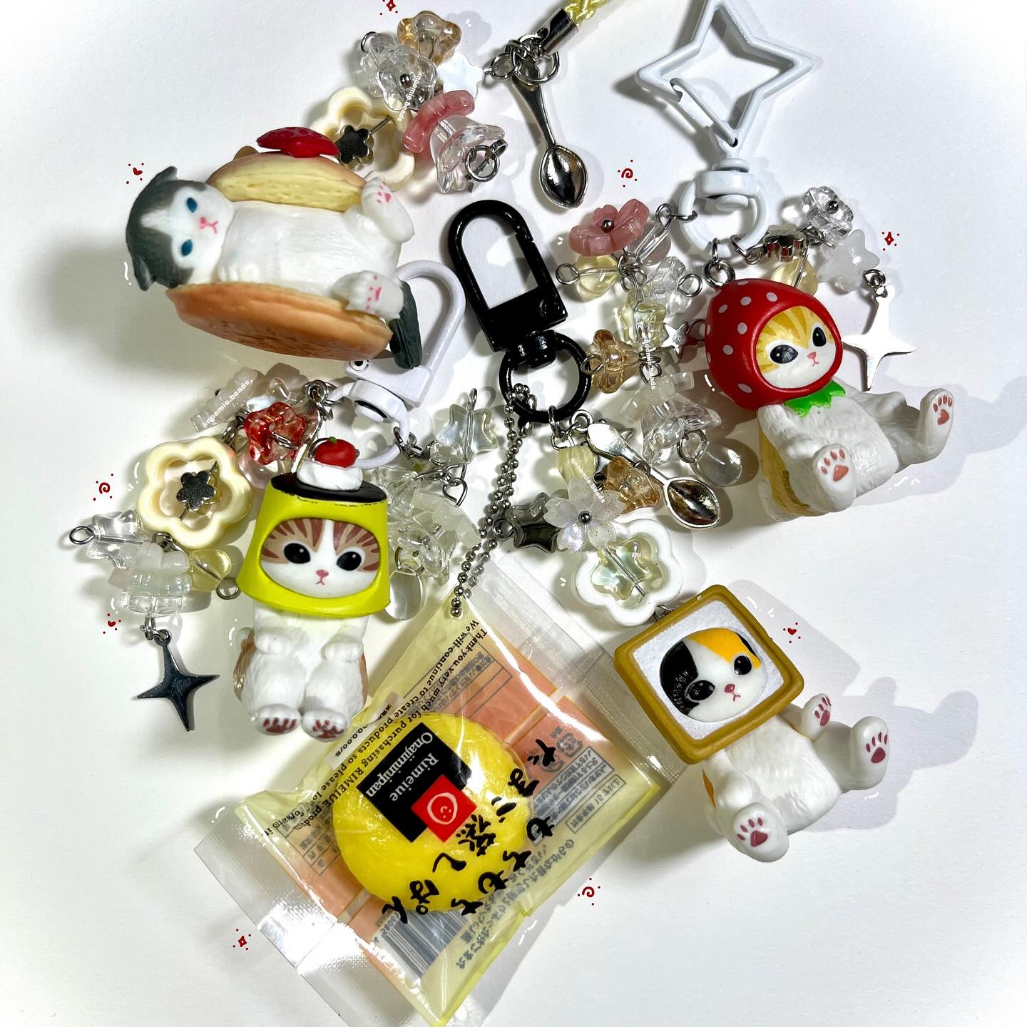 this image features a collection of cute and whimsical keychains and charms arranged on a white surface. the keychains are adorned with various cat figures, including a cat dressed as a pudding, a strawberry, a pancake, and even a cat with a tv head. other decorative elements include beads, flower charms, star charms, miniature spoons, and a black clasp. a japanese food packaging with text is also present among the charms. the overall aesthetic is playful and colorful.