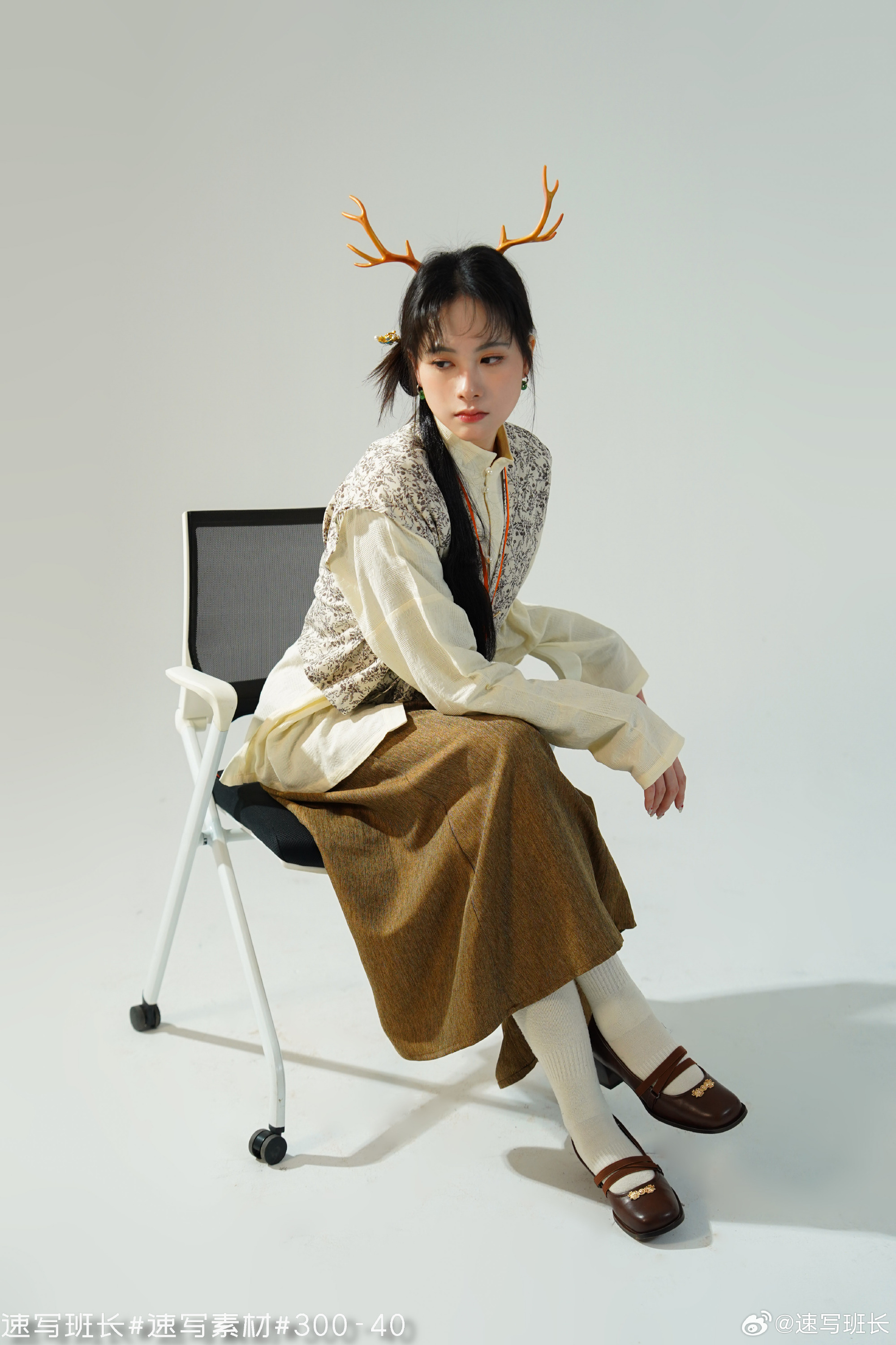a young woman with dark hair is wearing a pair of decorative antlers on her head. she is dressed in a stylish outfit consisting of a white shirt, a patterned vest, and a long brown skirt. she is seated on a black and white folding chair against a white background.  the image is welllit and features a simple composition.