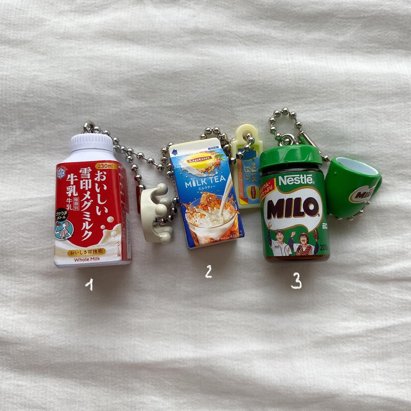the image shows a collection of drink miniature keychains. there's a milk bottle keychain labeled with japanese text, a lipton milk tea keychain, and a nestle milo can keychain with a small mug accessory. they all have silver ball chains and some have additional plastic connectors. the items are arranged on a white surface.