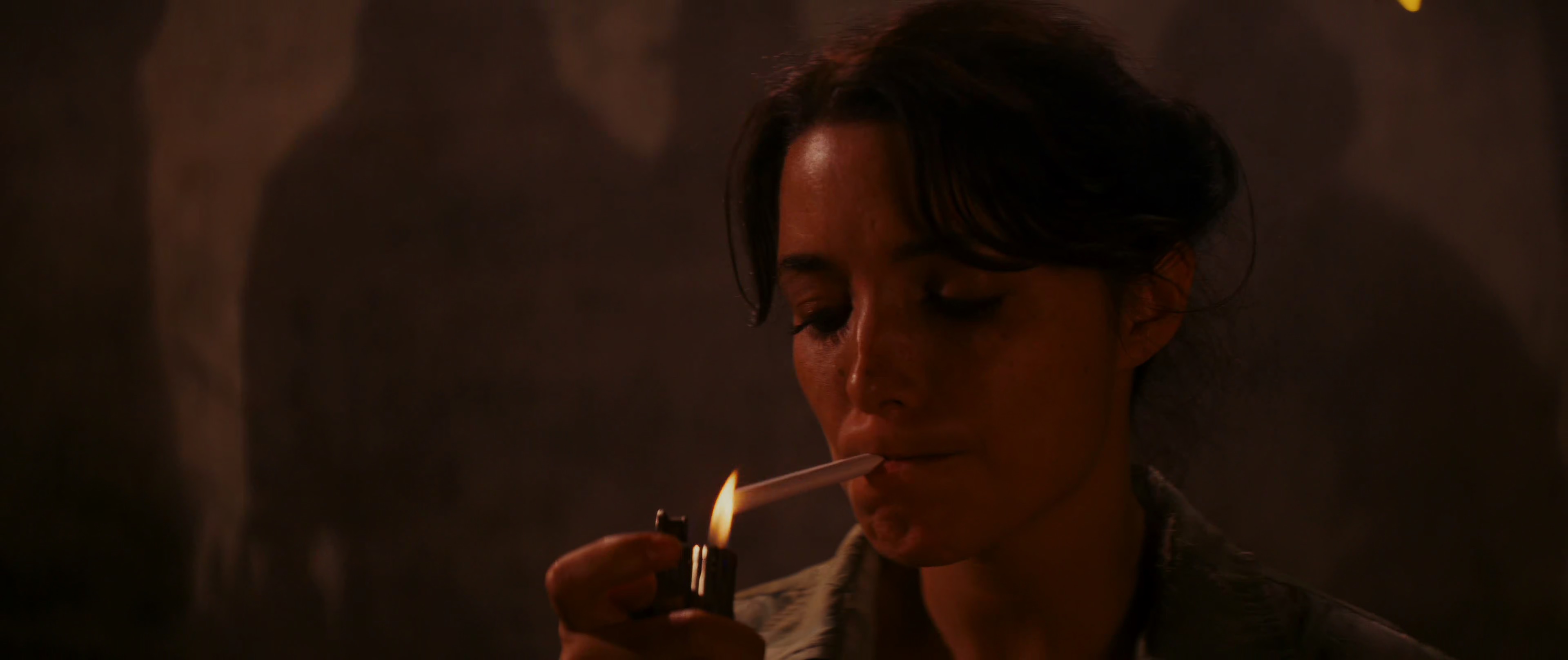 a closeup of a woman with long, dark hair, wearing a green shirt, lighting a cigarette with a lighter. she is looking directly at the camera, and her face is illuminated by the flame of the lighter. the background is dark and out of focus.