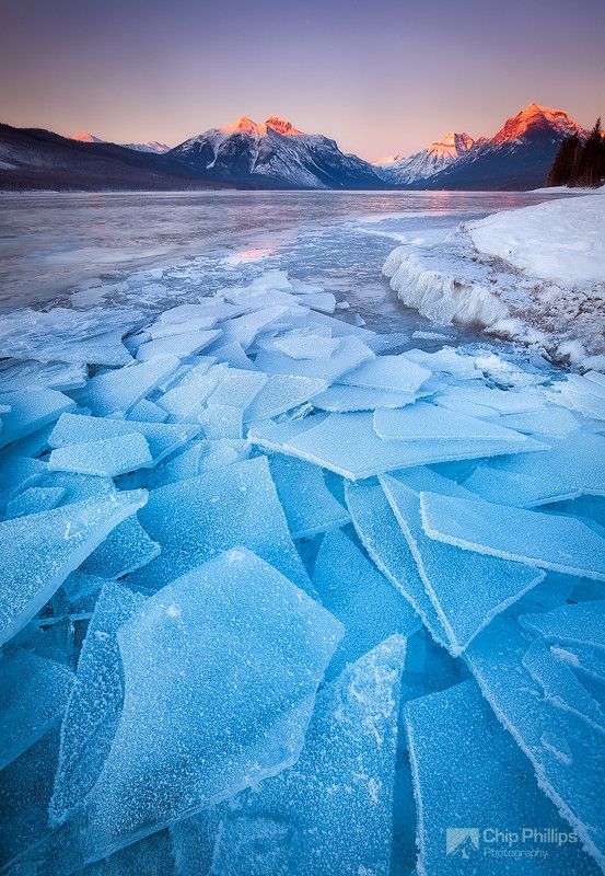 a wide shot of a frozen lake with mountains in the background. the lake is covered in a layer of ice that has broken into large, clear pieces. the mountains are snowcapped and the sky is a beautiful blend of pink, purple and blue. the sun is setting or rising, casting a warm glow over the landscape.