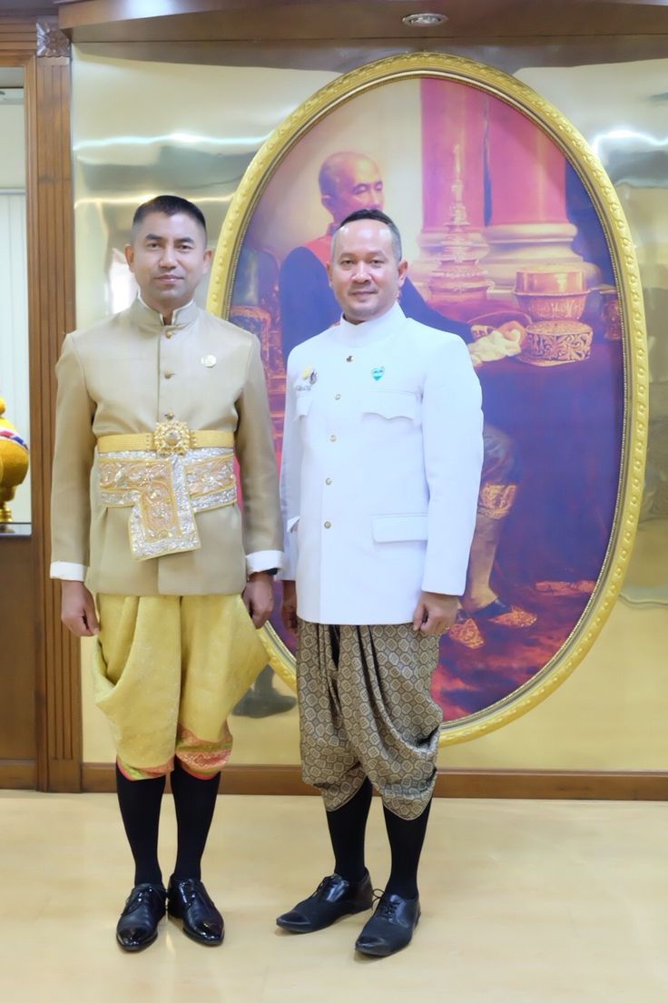 two men are standing in front of a painting. they are wearing formal thai traditional clothing. the man on the left is wearing a yellow longsleeve shirt and pants, while the man on the right is wearing a white longsleeve shirt with black pants. both men are smiling and looking at the camera. the background is a gold frame painting and a wooden wall.