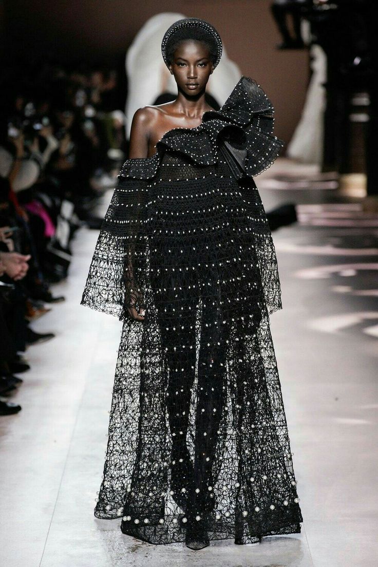 the image showcases a black model on a fashion runway, wearing a striking floorlength black gown. the dress features delicate lace or netting with pearllike embellishments throughout.  the neckline is offtheshoulder, and the bodice has a ruffled, slightly draped design.  the model is wearing a simple black headband. the overall style is elegant and highfashion, suggestive of a haute couture collection.  the background is blurred, focusing attention on the model and her attire.  the setting appears to be a professional fashion show, and the lighting is typical of such events.