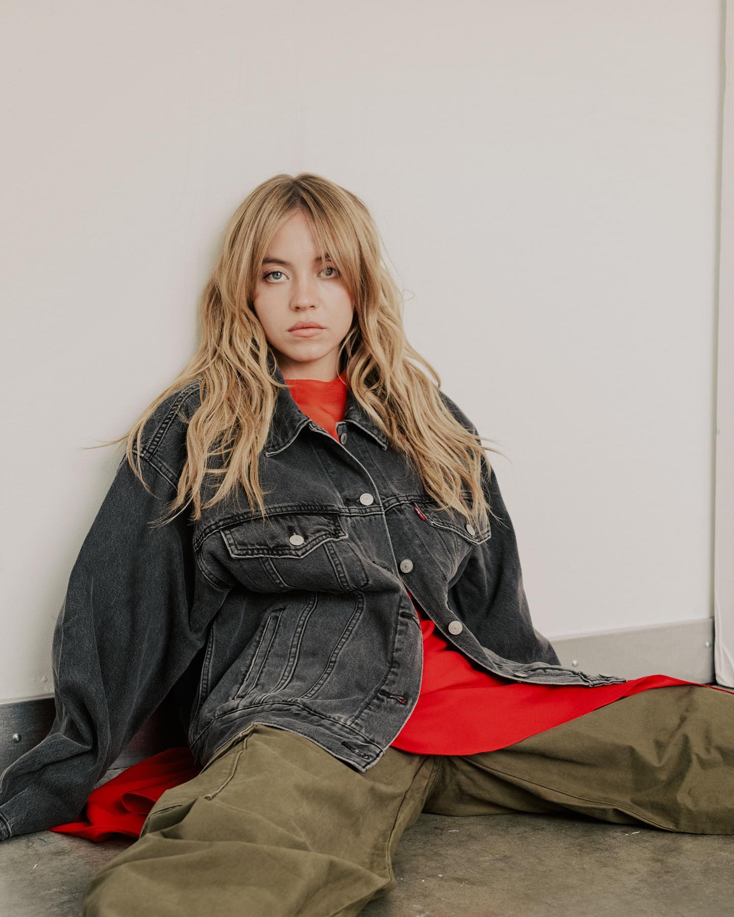 the image shows a young blonde woman with long wavy hair and bangs, sitting on the floor leaning against a white wall. she is wearing an oversized black denim jacket, a red tshirt, and olive green cargo pants. her expression is serious and slightly pensive.  the overall style is casual and relaxed with a hint of vintage or grunge influence. the setting appears to be a simple studio or indoor space. the photo is welllit and focuses on the model.