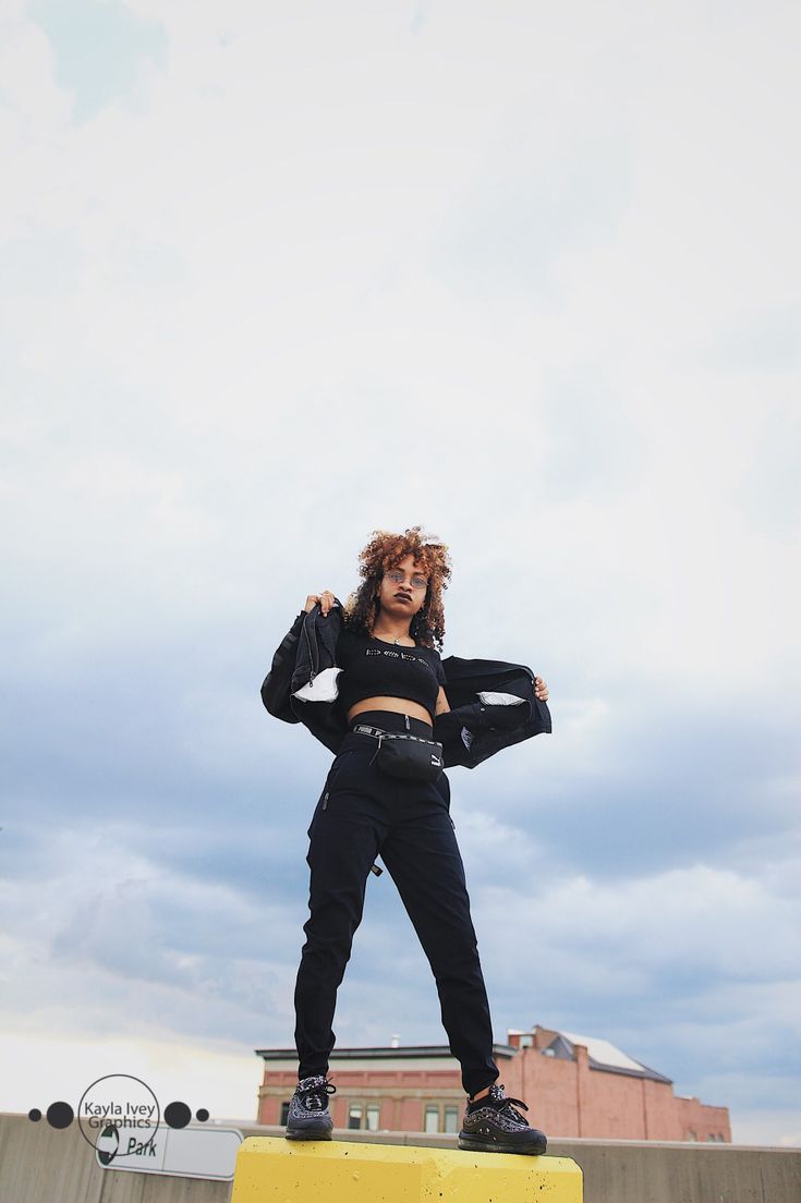 a young woman in a black crop top and pants, posing outdoors. she is wearing a black jacket over her shoulder, black sunglasses, and sneakers. she has her hair styled in a curly updo. she is standing in front of a city skyline. the sky is blue with white clouds.