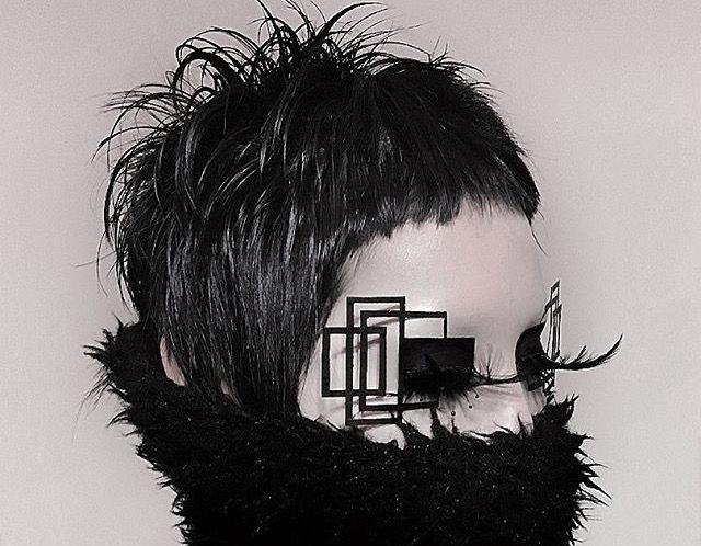 a woman with short black hair poses for a closeup portrait. she is wearing black makeup with geometric shapes and black fur clothing. the photo is in black and white, and the woman's expression is intense.