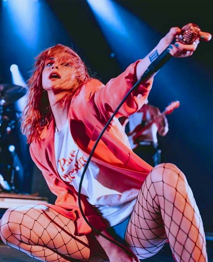 a vibrant image of hayley williams, the lead singer of paramore, performing live on stage. she is shown squatting, wearing a pink blazer, a white tshirt with writing, and fishnet stockings. she is holding a microphone with her right hand outstretched, seemingly engaging with the audience. blue stage lights illuminate her and the band behind her.