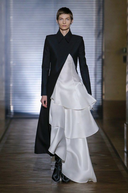 a model walks the runway during a fashion show, wearing a striking black coat with a white ruffled dress layered underneath. the model has short hair, neutral makeup, and black boots. the background features a blurred backdrop with a wooden floor and a sleek, modern setting. the model exudes confidence and sophistication, showcasing the latest trends in fashion.