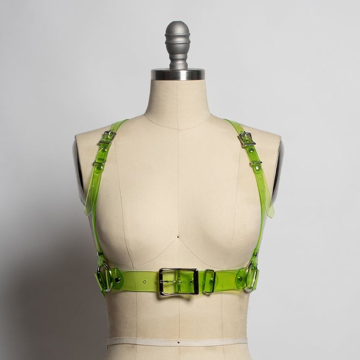 a green plastic harness with straps and buckles, worn by a mannequin. the harness is a fashion accessory that can be worn over clothing or as a statement piece. the harness is transparent, so the mannequin's body is visible through it. the harness has a beltlike design that cinches at the waist and has straps that go up the shoulders. the straps are adjustable so that the harness can be worn comfortably.