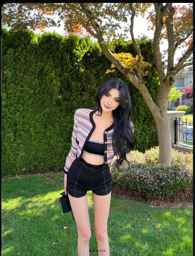 a young woman with long dark hair, wearing a pink and black sweater, black shorts, and a black bra, poses in a park. she is smiling and looking at the camera.