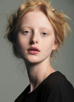 a portrait of a young woman with fair skin, blonde hair, and blue eyes. she is wearing a black shirt. her hair is styled in a loose, textured updo. she has freckles on her face and fair eyebrows. the background is a neutral grey.