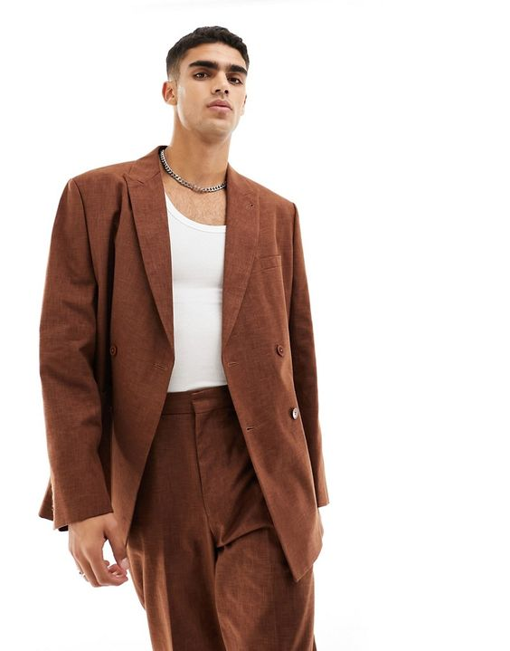 a young man standing in a brown double breasted suit with a white undershirt and a silver chain. the suit is relaxed and casual but stylish. he has a serious expression on his face.  the image is taken against a plain white background.