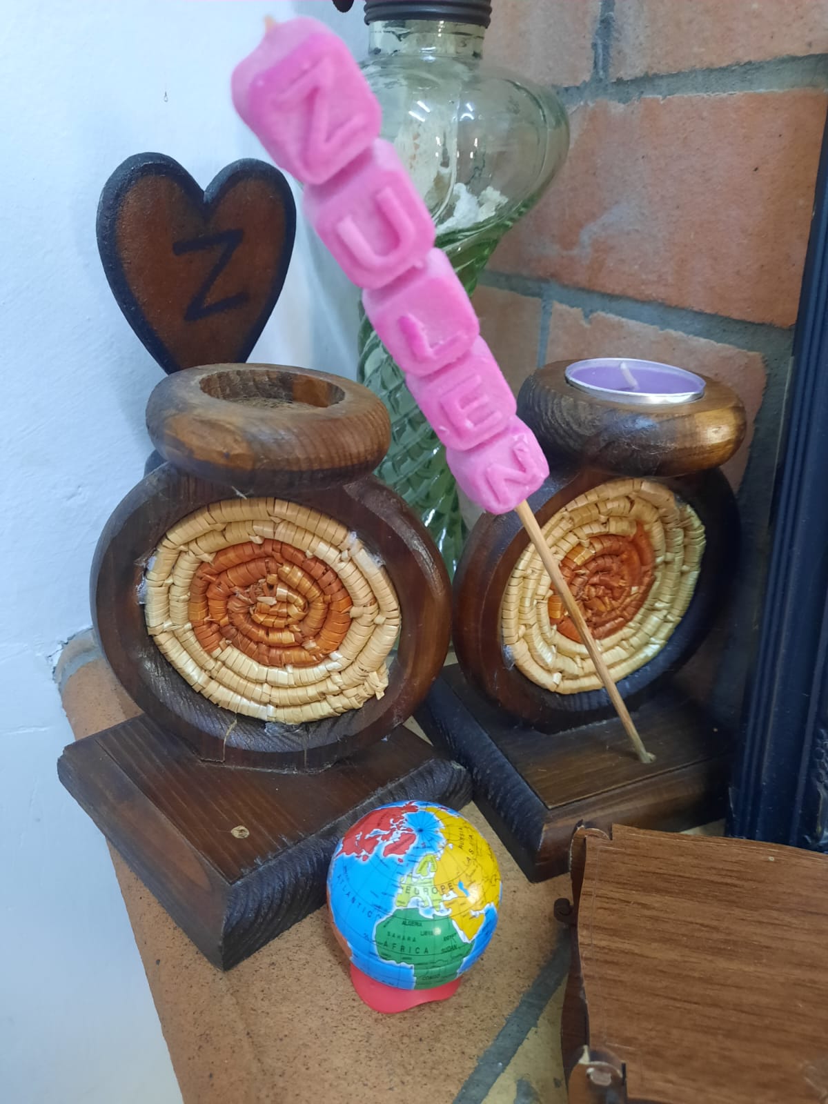 the image shows a variety of decorative objects, including two wooden candle holders with wicker inserts, a pink candle with the name "zuleen" spelled out in block letters, a small globe, a wooden heart with the letter "z", and a glass jar. the objects are arranged on a surface near a brick wall and a white wall.