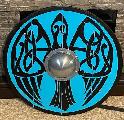 a round wooden shield painted with a stylized black raven on a light blue background. the raven's wings are decorated with black knotwork designs. the shield has a central metal boss.