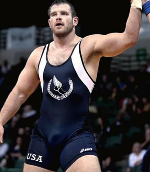 a muscular male wrestler wearing a usa wrestling singlet. the wrestler is in midaction and his arm is raised in victory. he is in a gymlike setting. the image is actionpacked.