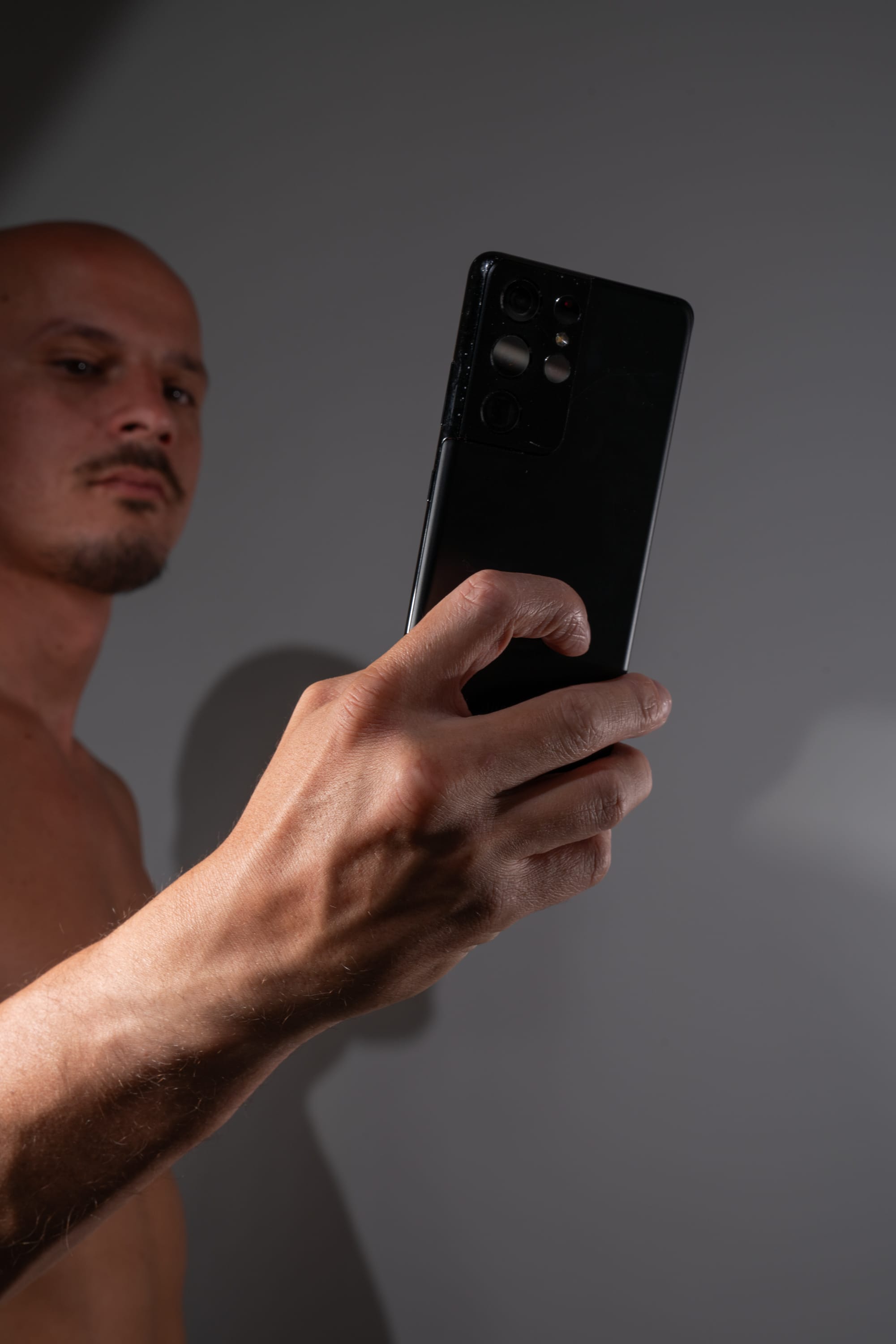 shirtless man holds a smartphone in front of him, taking a selfie. the focus is on the phone and his hand, while his face is slightly out of focus in the background. the lighting is dramatic, creating strong shadows across his body.