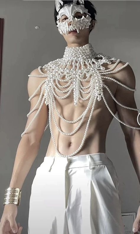 a man wearing a white mask, pearl necklace, white pants, and bracelet is standing shirtless. the mask is a bonelike design. the pearl necklace is multilayered. the pants are loosefitting and the bracelet is gold.