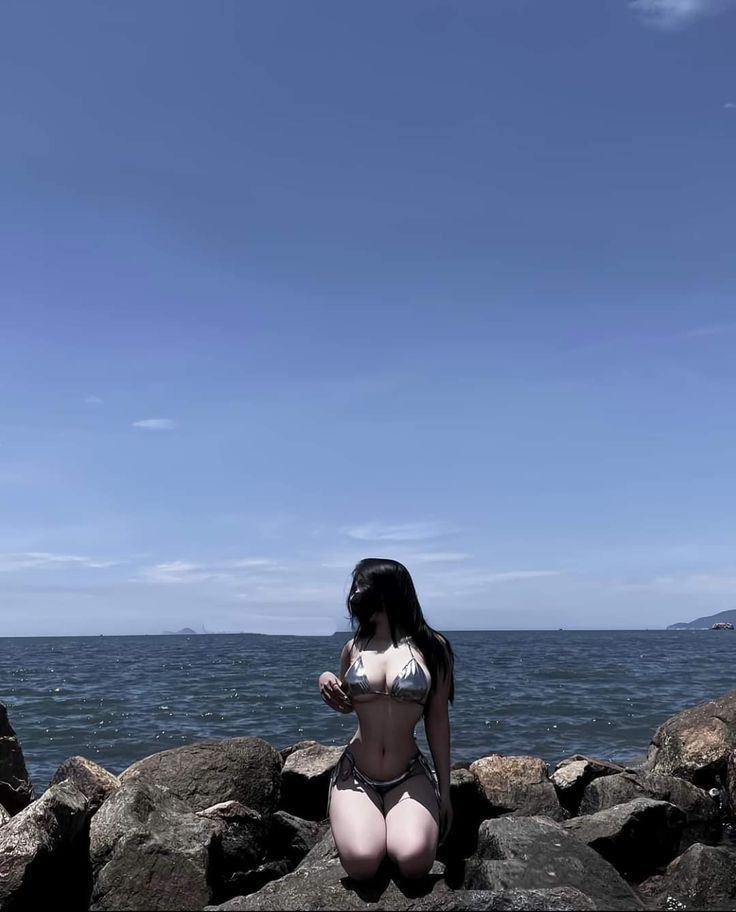 a woman wearing a silver bikini is kneeling on some rocks by the ocean on a sunny day.