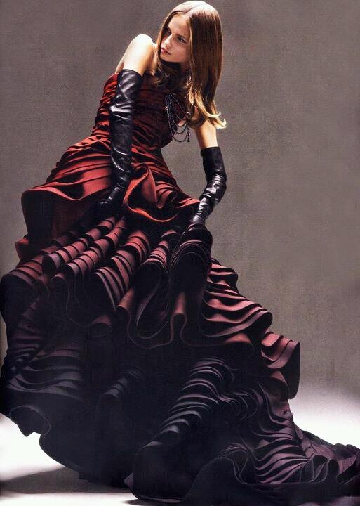 a woman in a long, flowing red and black dress with ruffle details and black gloves is standing in a studio setting. she is looking down and her expression is serious. she is wearing a long dress with a ruffled skirt that is gathered at the waist and a bodice that is fitted. she is wearing black leather gloves that reach to her elbows. the background is a plain, gray wall. the lighting is soft and diffused. the image is a highfashion portrait.