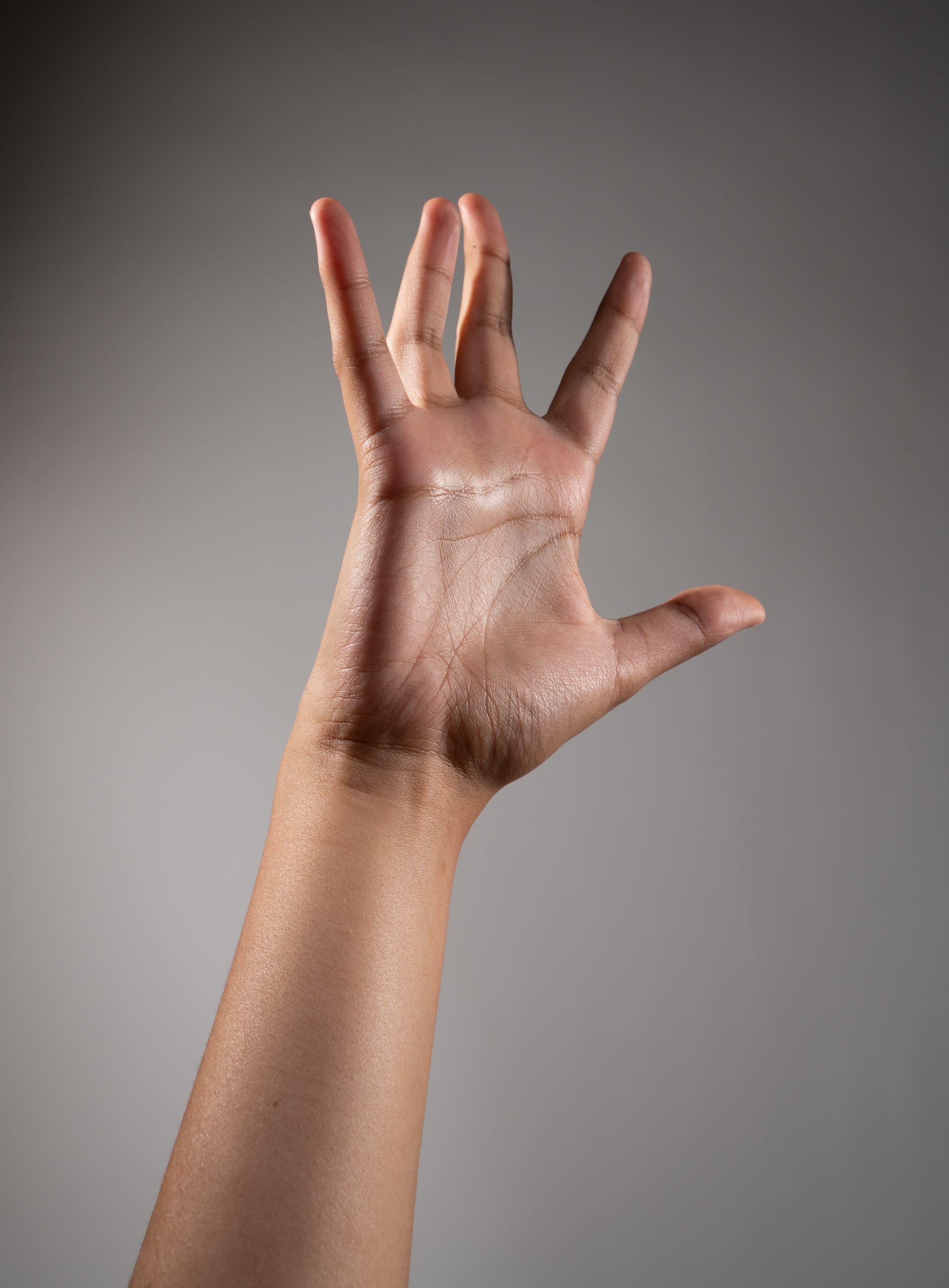 the image shows a woman's left hand and arm extended upwards against a gray background. the hand is open, with the fingers slightly spread apart. the skin of the hand and arm is brown and smooth, with highlights from the studio lighting. the gray background is even and unobtrusive, drawing attention to the hand.