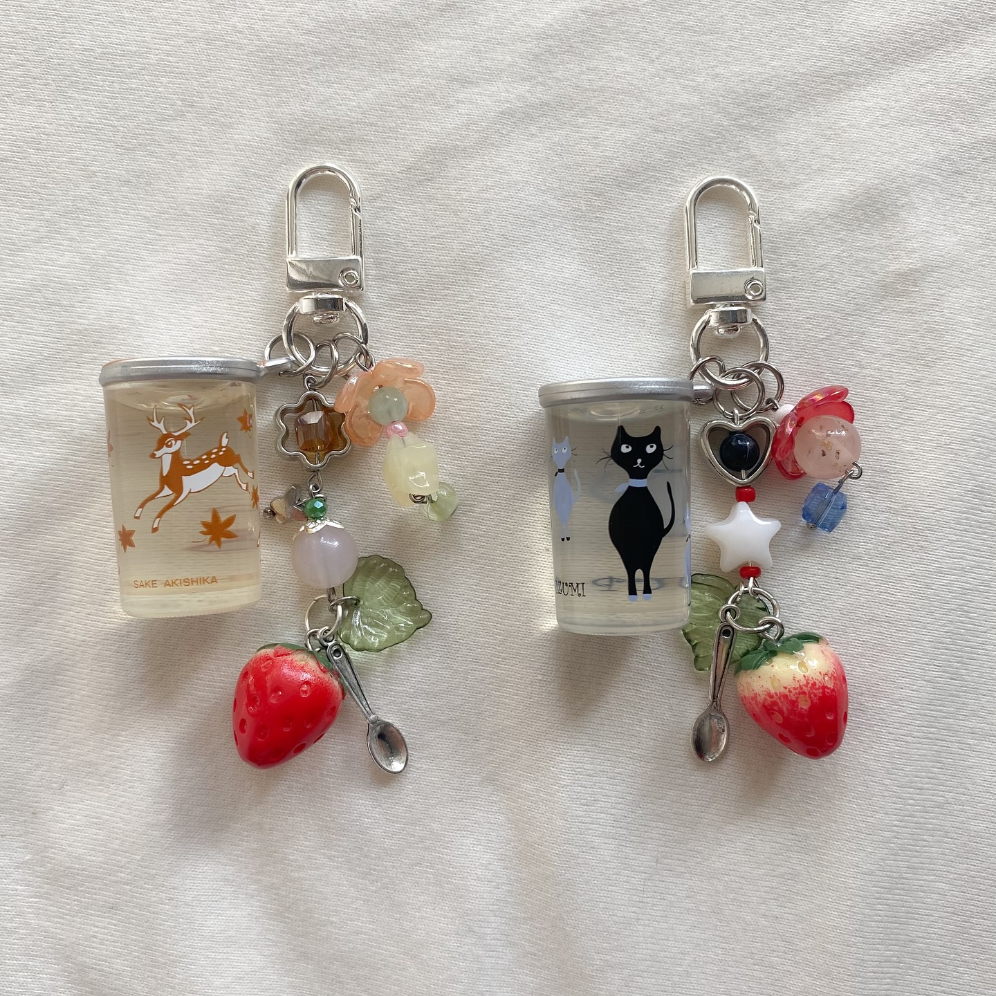 two handmade keychains lie on a white surface. each keychain features a silver clip attached to a small plastic cupshaped charm with different designs. one cup has a deer motif, and the other features a black cat. dangling from each cup are a variety of beads and charms, including a strawberry, a spoon, flower beads, a leaf, a star, a heart, and glass beads. the keychains have a whimsical and personalized look.