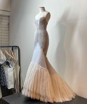 a long, creamcolored wedding dress is displayed on a mannequin. the dress has a strapless bodice with a sweetheart neckline. it flares out at the knees into a mermaidstyle skirt with layers of tulle, creating a voluminous train. a garment rack with several clothes hangs on the left. the mannequin and dress are set on a dark surface with a plain white wall behind it.