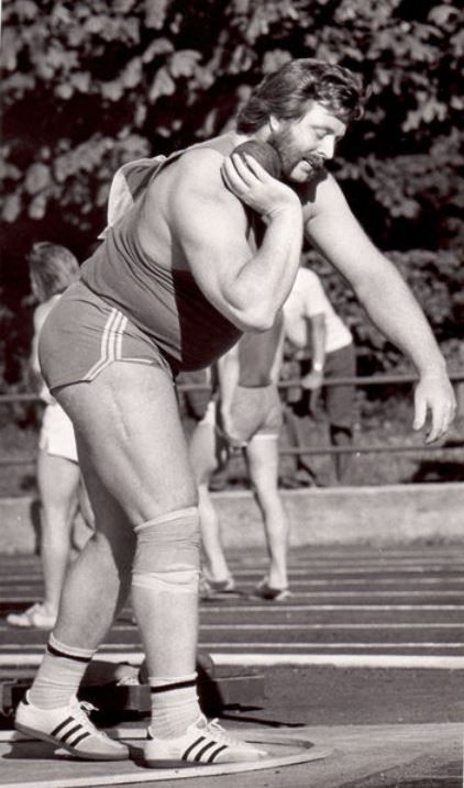 a black and white image of a shot putter in the middle of competition. he's holding the shot put near his neck, ready to throw. he's wearing athletic clothes and adidas shoes. there are other athletes in the background. the image appears to be taken outdoors, with trees in the background.