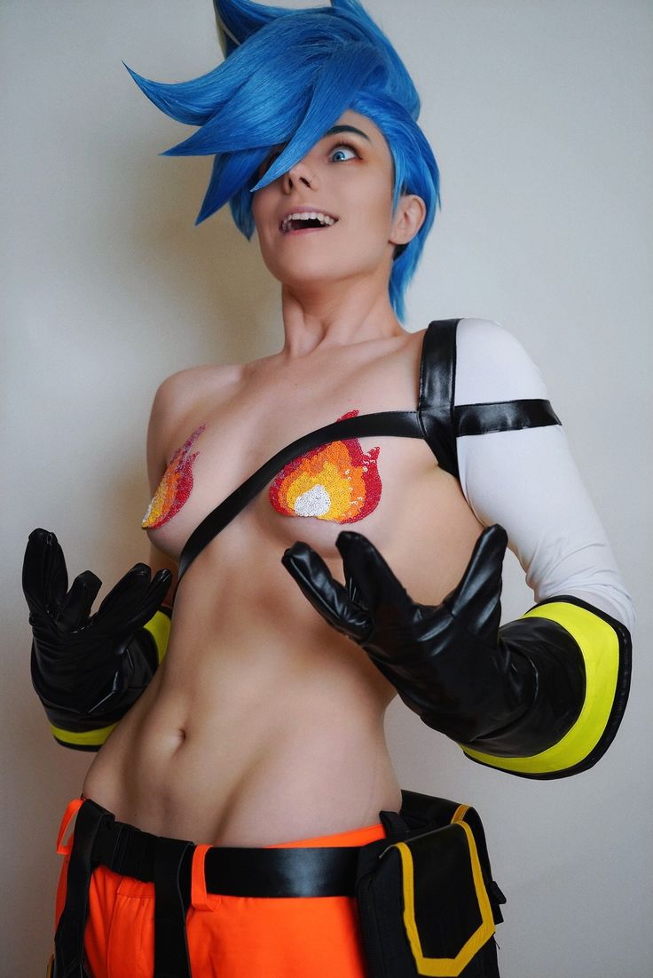 a cosplayer dressed as a genderbent version of yoko littner from gurren lagann poses for a photo. the cosplayer has a blue wig, wears flame pasties, gloves, and orange pants. they are looking up with a surprised expression.
