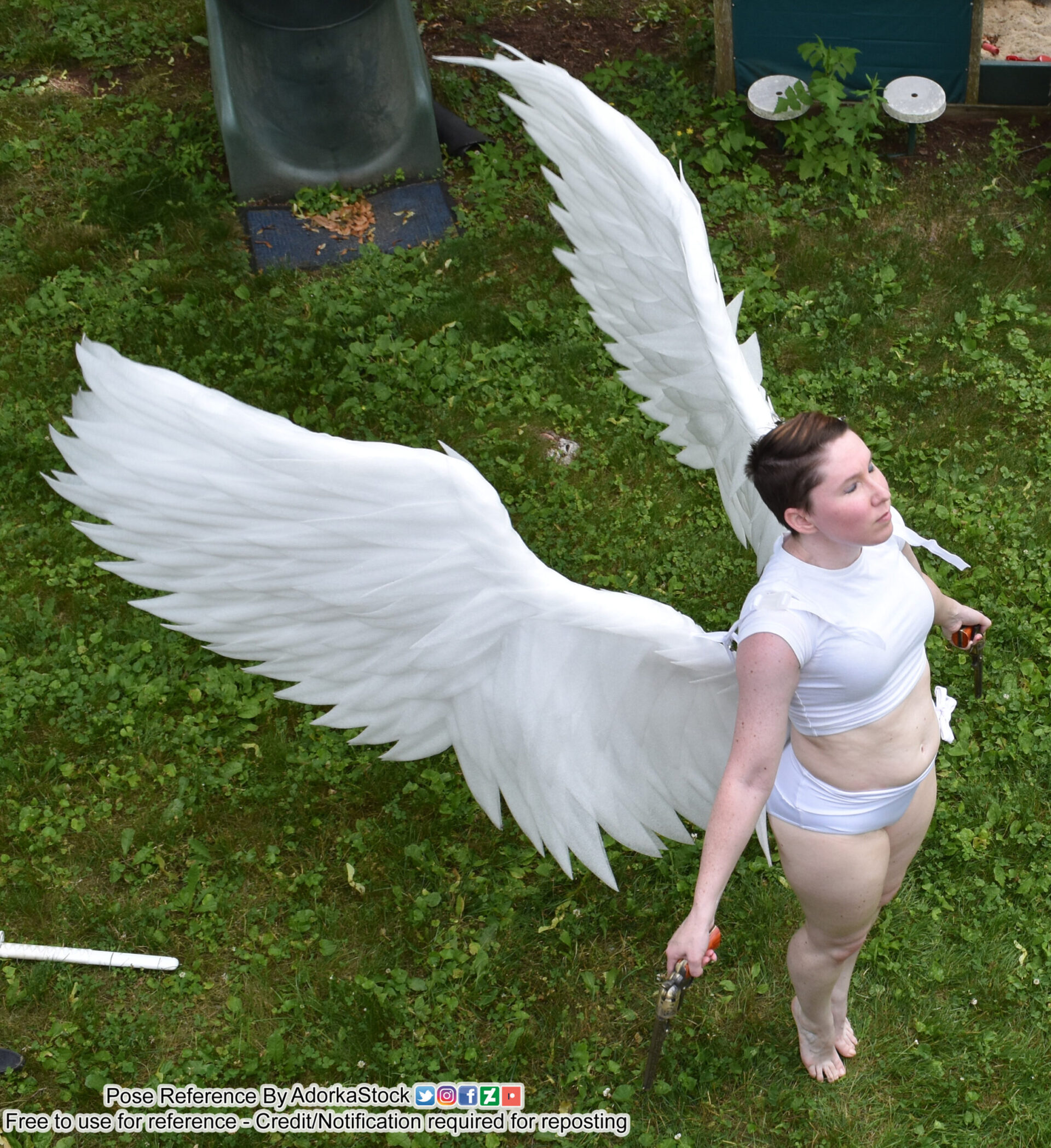 a woman in a white angel costume, stands in a grassy area with a rope in her hand. she's wearing a white top and white shorts. she's looking upward with a serious expression. she has white wings on her back. the wings are feathered and look to be made from a soft material. she's standing on a patch of green grass. the area is overgrown with small green plants. there is a small green plastic toy near her feet. the image is taken from a low angle and has a shallow depth of field. the wings are in focus and she is partially in focus.  the grassy area is blurred in the background. the light is bright and casts shadows in the grass.