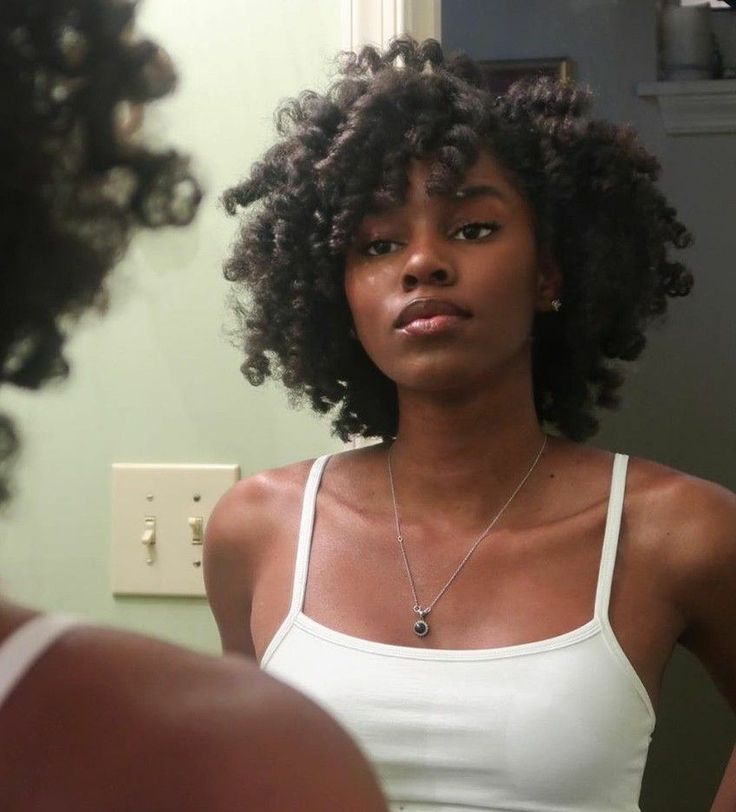 a woman with curly hair stands in front of a mirror, wearing a white tank top and a necklace with a pendant. her reflection can be seen in the mirror, and a light switch is visible on the wall. the setting appears to be a bathroom.
