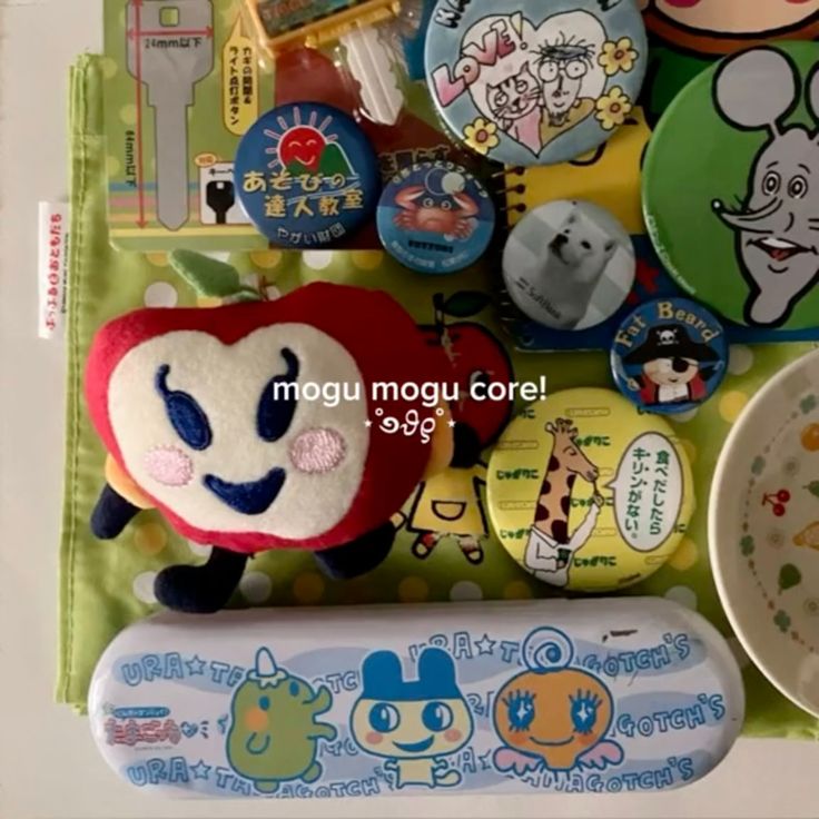 the image contains a variety of items including a red plush toy with a happy face, several round buttons with different designs, a key guide, and a pencil case with tamagotchi characters on it. the items are arranged on a green and patterned surface.