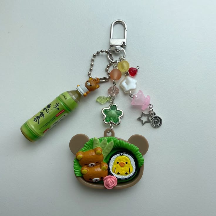 a closeup shot of a kawaii keychain featuring a miniature green tea bottle, various beads and charms, and a bearshaped piece containing a tiny bear figure and a chick, decorated with a flower.
