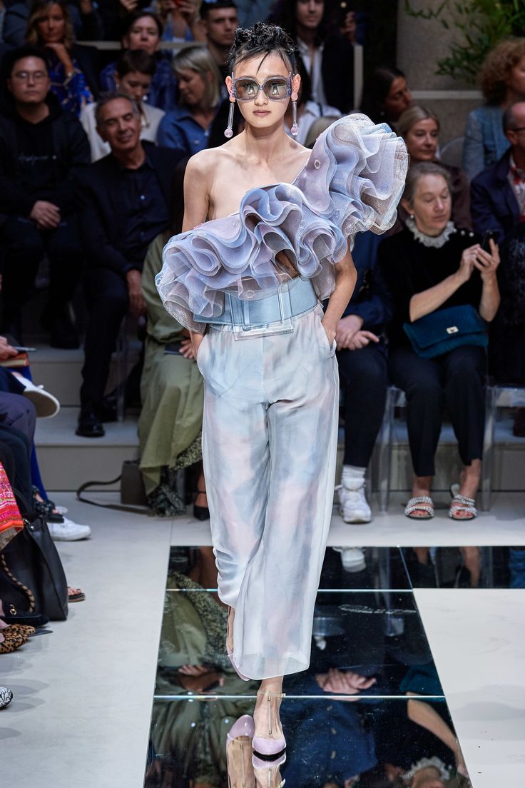 a beautiful woman walks the runway in a sheer dress with ruffles,  she is wearing white pants and silver sandals. she has a bright smile and her hair is pulled back in a ponytail with loose strands around her face.  she is surrounded by an audience of people wearing their best clothes.  the background is a black and white checkered floor.