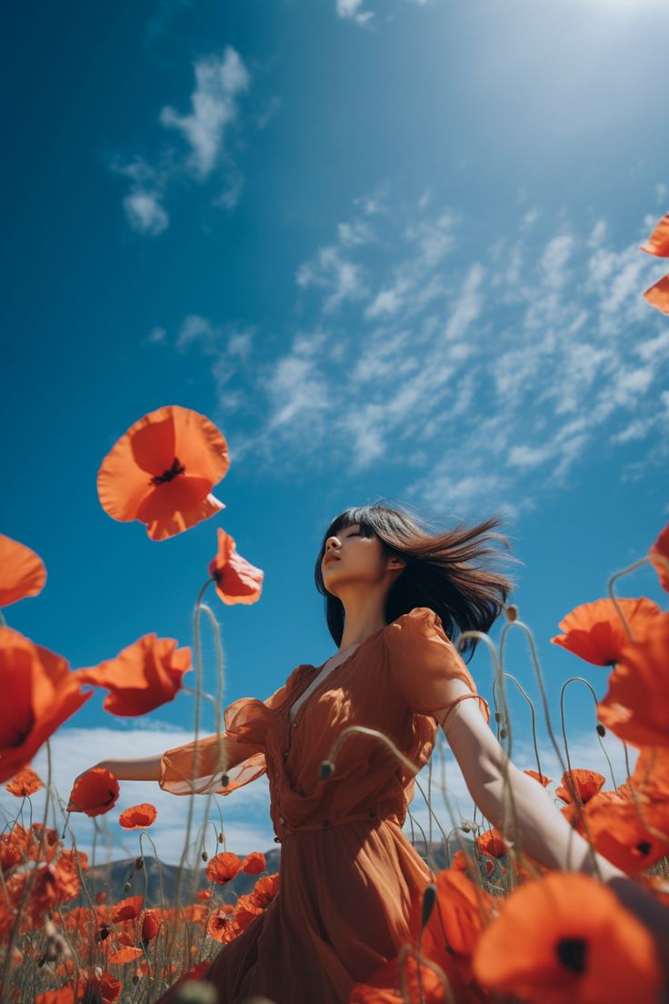 the image is a vibrant, lowangle shot of a young woman in a flowing orange dress standing amidst a field of bright red poppies. the sun is shining, and the sky is a clear, brilliant blue with fluffy white clouds. the woman's hair is blowing in the wind, her eyes are closed, and she appears peaceful and content. the composition emphasizes the vastness of the poppy field and the woman's immersion within its beauty.  the overall mood is one of serenity and joy, capturing a moment of connection between the woman and the natural world.