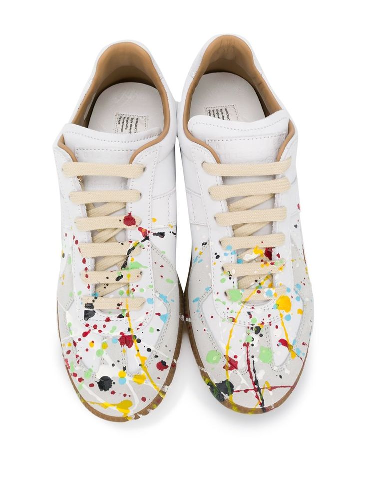a pair of white leather maison margiela sneakers with a paint splatter design. the sneakers have beige laces and a gum sole. they are shown from above, and the paint splatters are visible on the toe and sides of the shoes.