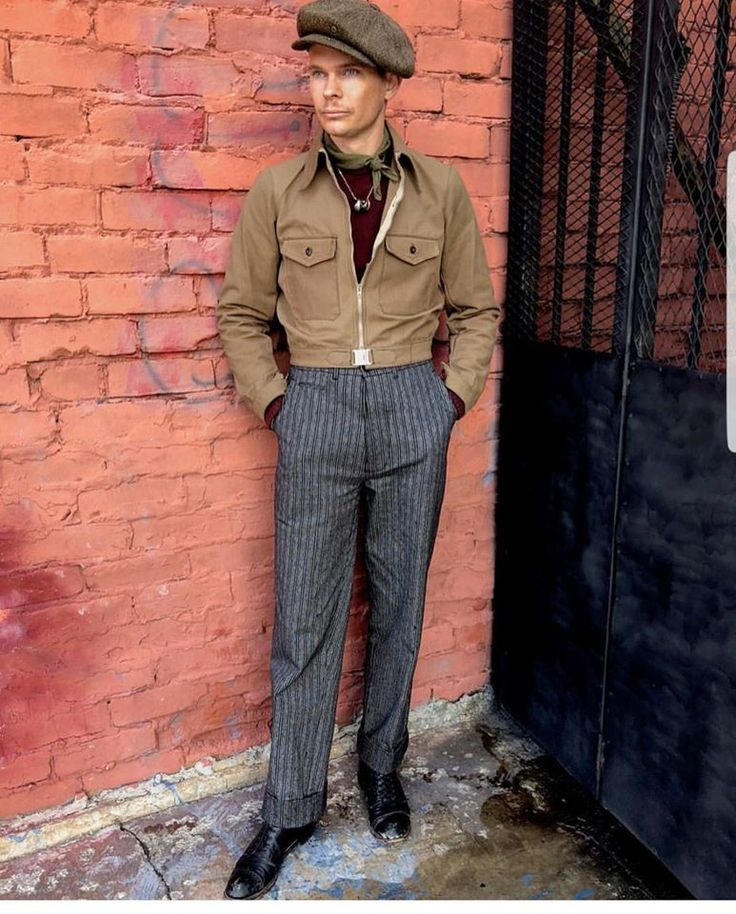 the image shows a man standing against a red brick wall. he is wearing a vintageinspired outfit that includes a beige cropped jacket, grey pinstripe highwaisted trousers, black oxfords, a newsboy cap, and a burgundy turtleneck. the style evokes a 1940s or 1950s aesthetic.  the man is looking directly at the camera. the background includes a section of a metal gate. the overall aesthetic is sharp and stylish.
