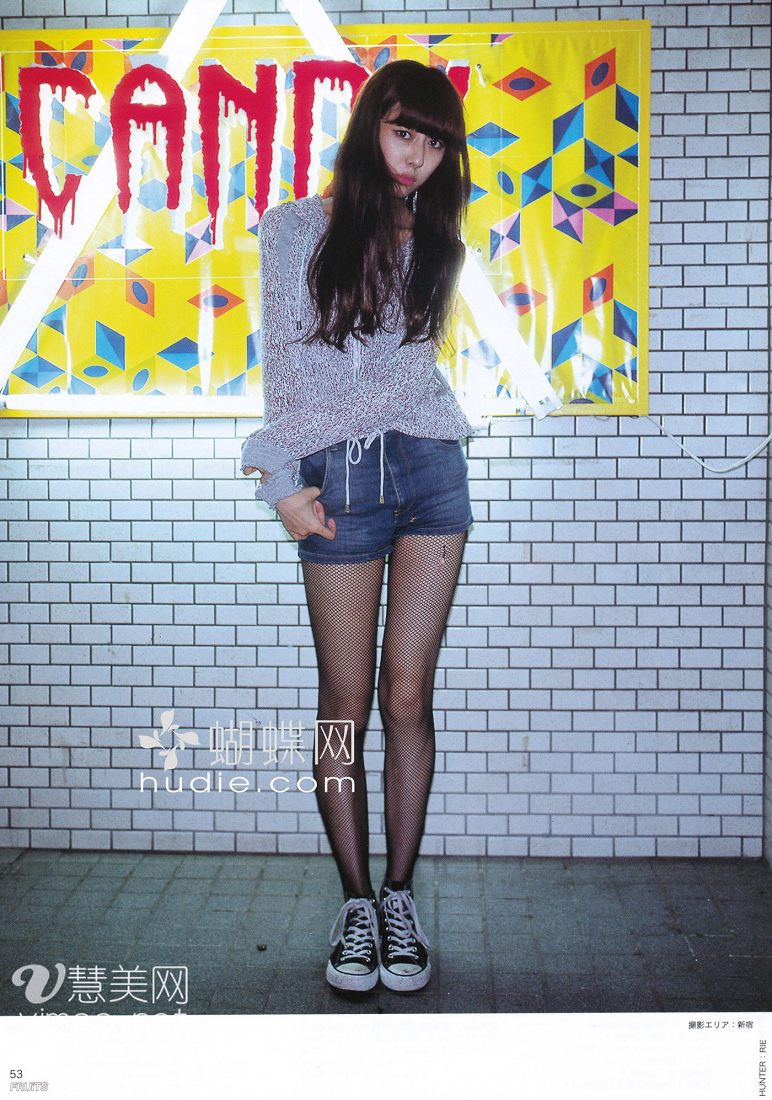 a young woman with long, dark hair and bangs is posing in front of a graffiticovered wall. she is wearing a gray sweater, denim shorts, black fishnet stockings, and black and white converse sneakers. she has a confident and stylish demeanor. the lighting is bright and vibrant, highlighting her outfit and the colorful graffiti behind her.