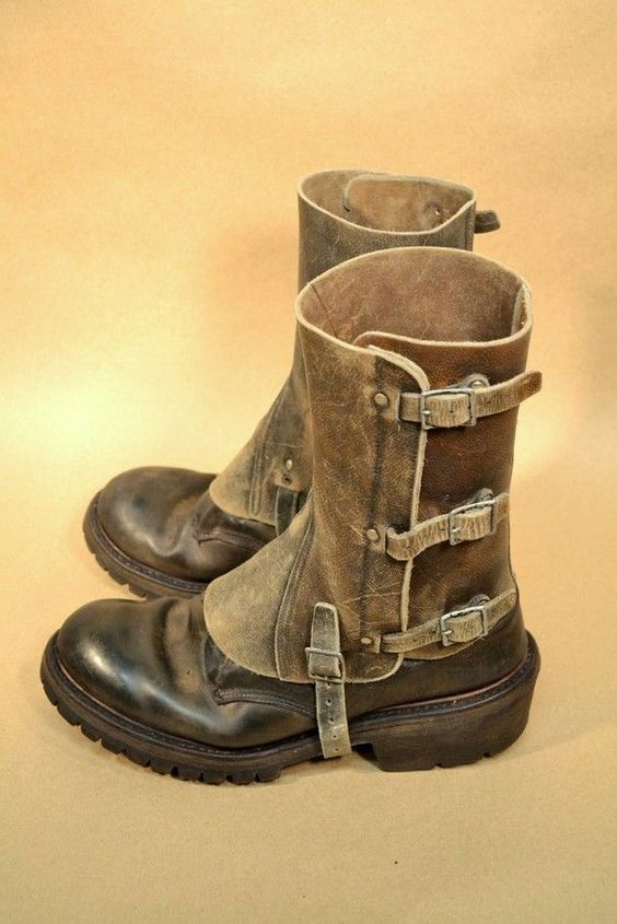 a pair of brown leather motorcycle boots with buckles and straps, worn and distressed. the boots are sitting on a light brown background. they have a worn vintage look, with visible scratches and wear. the buckles are all secured and the boots are ready to be worn.