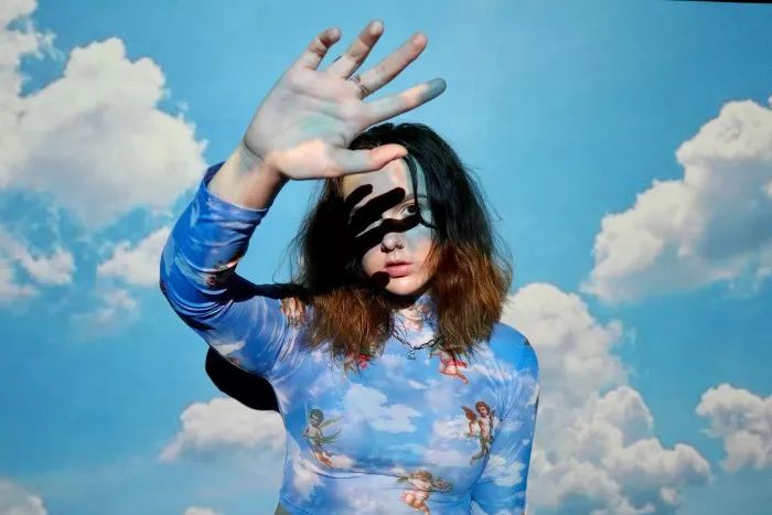 a young woman with long brown hair is wearing a blue shirt with clouds and angels printed on it. she is posing with her hand covering her face and looking at the camera. the background is a blue sky with clouds.
