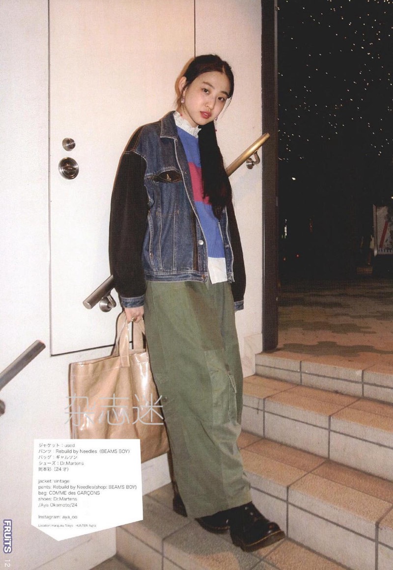 a young woman poses on an urban street at night. she is wearing a vintageinspired outfit consisting of an oversized denim jacket, a colorful sweater, olive green cargo pants, and black doc martens boots. she carries a large, beige tote bag. the lighting is low and atmospheric, highlighting the grungy streetwear aesthetic of her look.