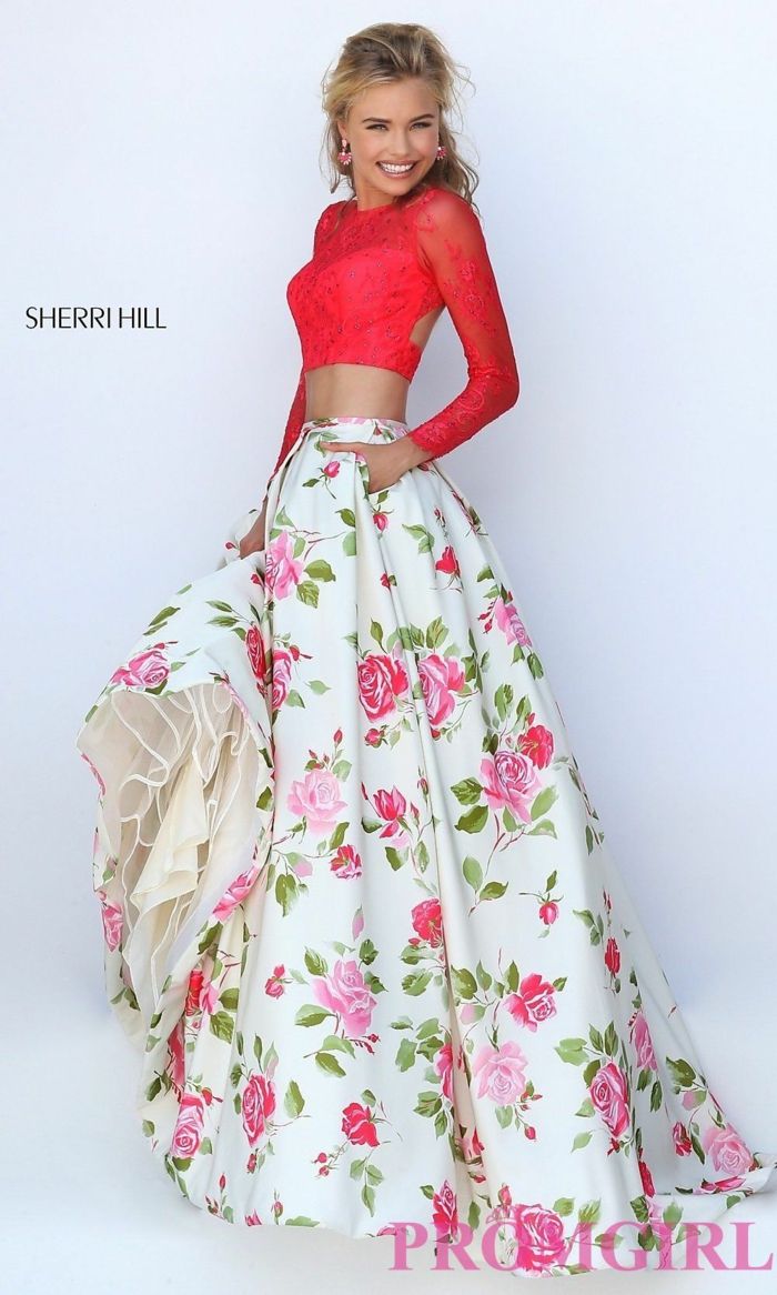 a woman is standing wearing a twopiece gown. the top is a longsleeved red crop top with lace details. the skirt is a white floral pattern with pink and red roses and green leaves. the skirt is full and flows to the floor. the woman is holding up the skirt on one side, revealing a lightercolored lining. the dress has the sherri hill logo in the background. the bottom right corner of the image has the progirl logo.