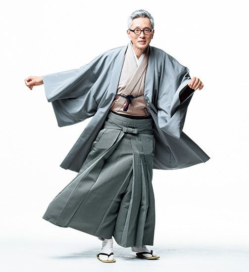 a senior man in a grey kimono and hakama, posing and smiling.  he is wearing a traditional japanese outfit.  the background is white.