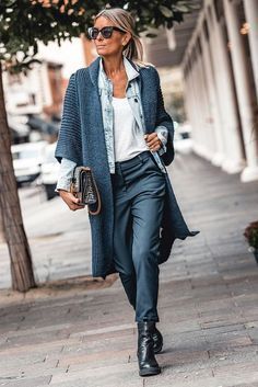 a woman wearing a stylish outfit is walking on a city street. she is wearing a blue cardigan, blue jeans, black pants, black boots, and sunglasses. she is carrying a brown bag.