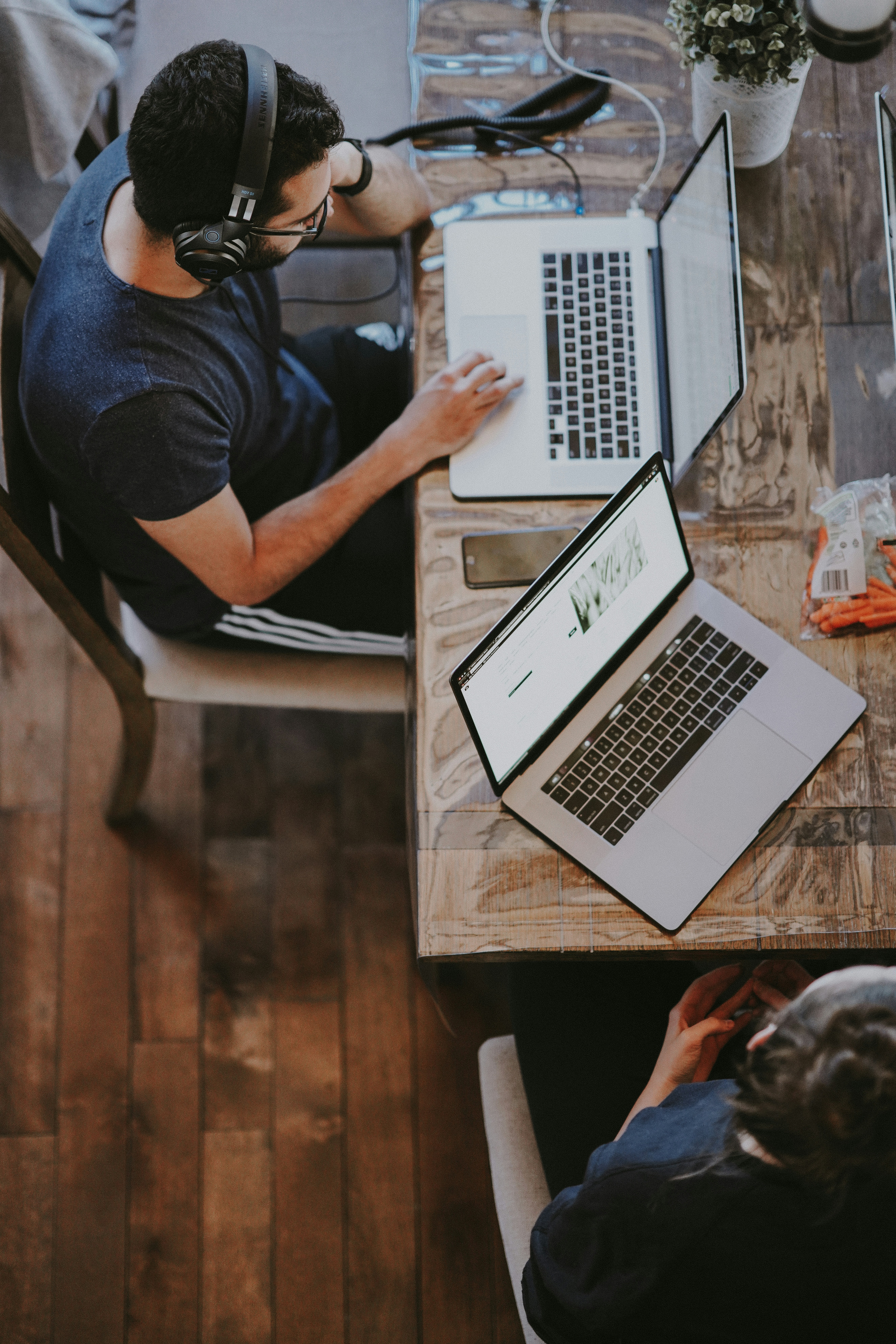 the image showcases an aerial view of two individuals working from home, seated at a wooden table. the man, wearing headphones, is engrossed in his work on a laptop while the woman sits opposite him, her laptop open and hands resting on her lap. the scene exudes a casual yet productive atmosphere, illuminated by natural light streaming in from the side, highlighting the wooden floor and the warm tones of the furniture.